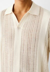 Polo de punto de color beige claro que presenta un diseño texturizado y perforado con franjas verticales y un cuello clásico.