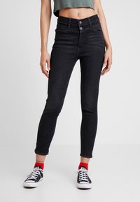 Jeans ajustados negros de cintura alta, con cinco bolsillos y detalles sutilmente desgastados. Combinados con zapatillas negras y calcetines rojos por el tobillo.