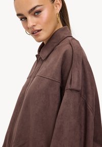 Veste-chemise en faux daim marron avec un col structuré, des épaules tombantes et un détail de passepoil subtil, mettant en valeur une texture douce et une coupe décontractée.