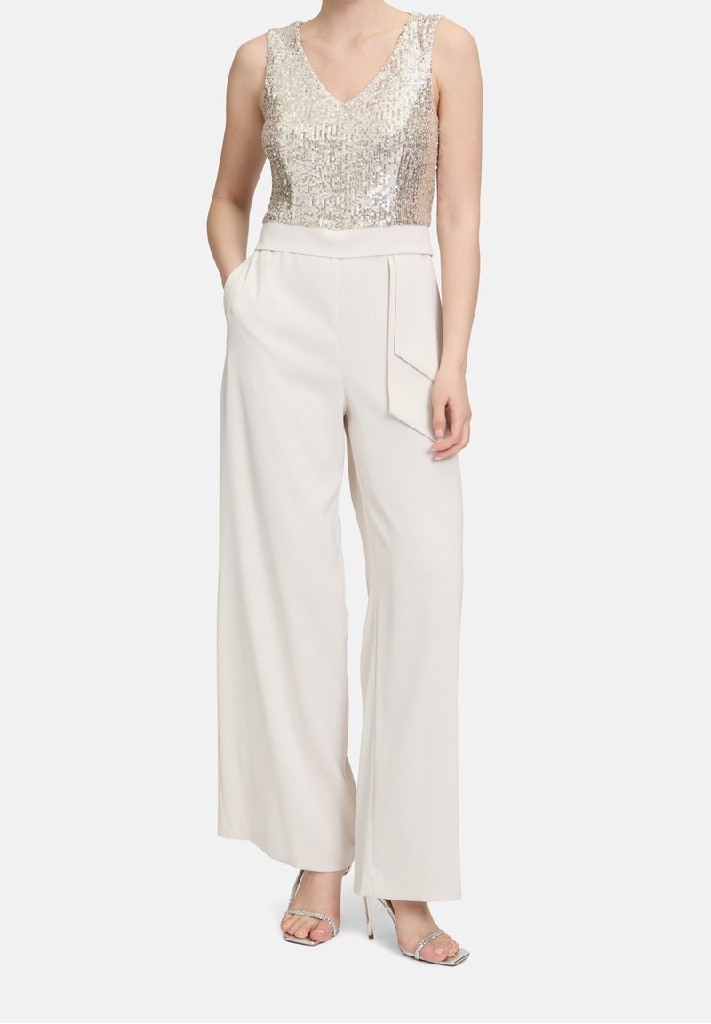Haut sans manches à paillettes dorées, associé à un pantalon blanc à jambes larges avec poches latérales et détail plissé à l'avant. Sandales à talons argentées.