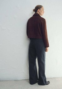 Braune, kürzere Wildlederjacke mit Kragen, kombiniert mit hoch taillierten, gerade geschnittenen schwarzen Jeans und schwarzen Schuhen. Neutraler Hintergrund.