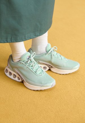 Person wearing white socks and light green Nike sneakers with visible Air cushioning, standing on a yellow surface.