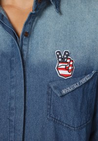 Camisa de mezclilla en azul claro con botones marrones. Presenta un símbolo de paz bordado en rojo, blanco y azul y un parche que dice "ABSOLUTAMENTE PERFECTO".