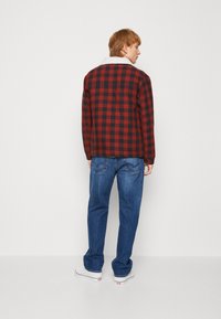 Camisa jaqueta xadrez em vermelho e preto com um colarinho de pelúcia branca, vestida sobre jeans azuis e sapatilhas brancas, vista pelas costas.