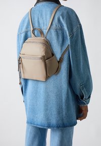 Osoba oblečená v světle modré džínové bundě a kalhotách, s malým béžovým koženým batohem na zádech, na jednobarevném pozadí.