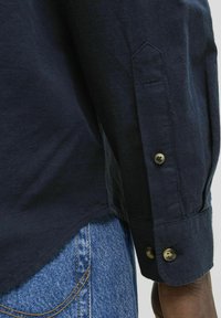Marineblauw overhemd met lange mouwen, gemaakt van een gestructureerde stof, met knopen bij de manchetten en een iets gebogen zoom, gecombineerd met lichtblauwe denimspijkerbroeken.