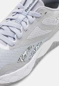 Světle šedá sportovní obuv s použitím síťoviny a suede materiálů, texturovaný povrch, bílá podrážka a černý vzor na bočním panelu.