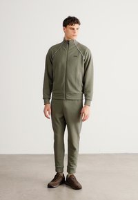 Αθλητική φόρμα σε ελαιί πράσινο, φτιαγμένη από μαλακό ύφασμα. Διαθέτει μπουφάν με φερμουάρ και αντίθετη τρέσα, καθώς και παντελόνι στενό με ελαστικά τελειώματα.