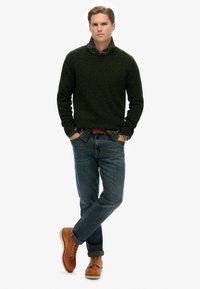 Suéter de punto verde con manchas, camisa de cuadros debajo, jeans de mezclilla azules y botas de cuero marrón claro con diseño de cordones. Atuendo simple y casual.