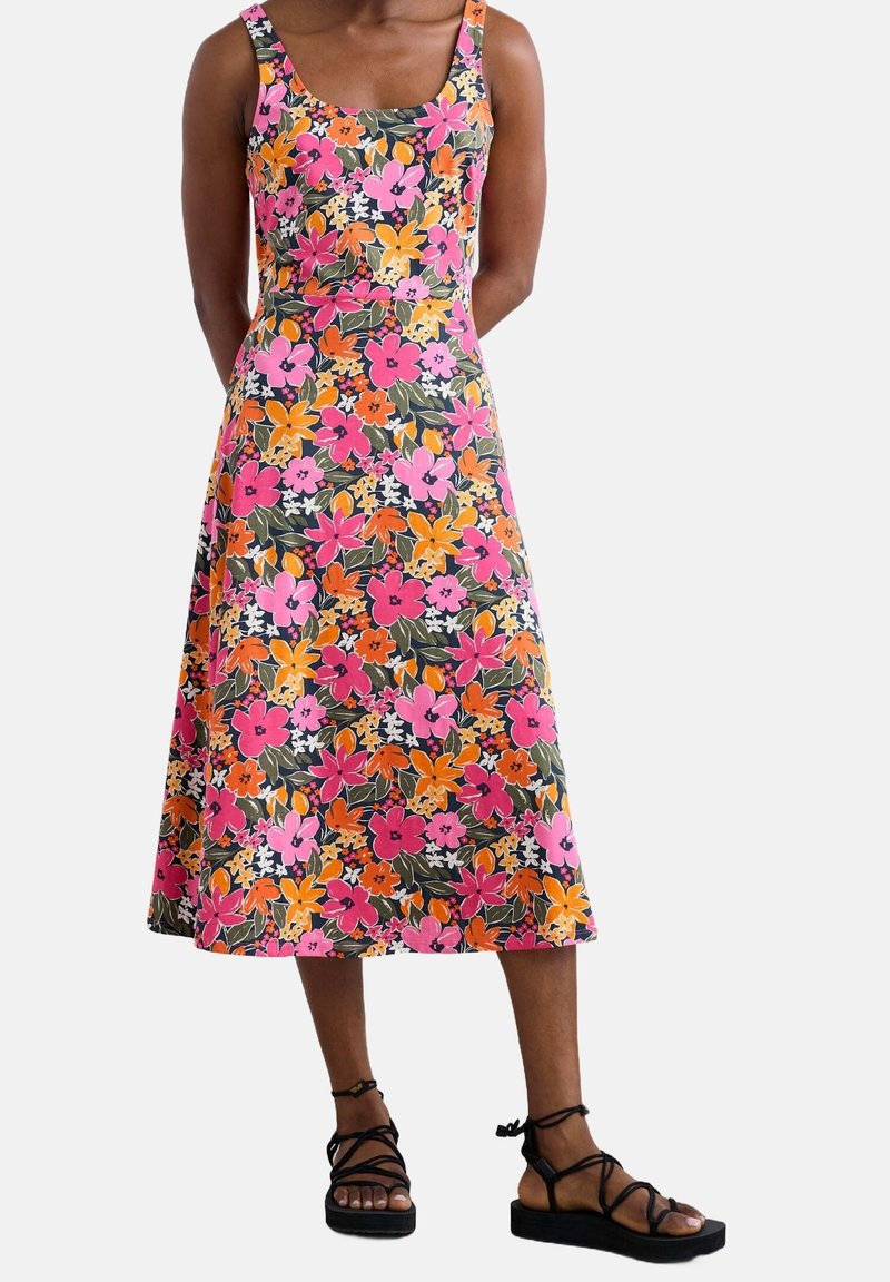 Femme portant une robe midi sans manches avec un imprimé floral rose vif, orange et blanc et des sandales noires à brides sur un fond uni.