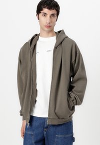 Olive green zip-up hoodie with a relaxed fit, large hood, and side pockets, layered over a white t-shirt with light blue graphic details.