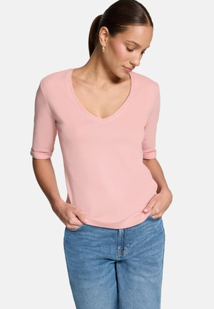 Žena s hnedými vlasmi v cope, ktorá nosí zlaté kruhové náušnice, ružové tričko s V výstrihom a vyhrnutými rukávmi a svetlomodré džínsy, ruky v kapsách.