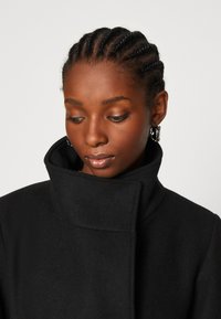 Cappotto di lana nera con colletto alto, texture liscia e design minimalista. La modella indossa orecchini d'argento, mostrando un'espressione neutra.