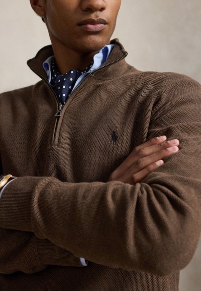 Άνδρας με σταυρωμένα χέρια, που φοράει καφέ μάλλινο πλεκτό ζιπ-απ πουλόβερ, μπλε πουκάμισο και σκούρο μπλε φουλάρι με πουά, με ρολόι ορατό στον καρπό.