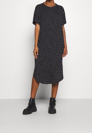 Sort knælængde kjole med hvide polkaprikker, korte ærmer, afrundet kant, båret med sorte ankelstøvler med spænde på en stående person.