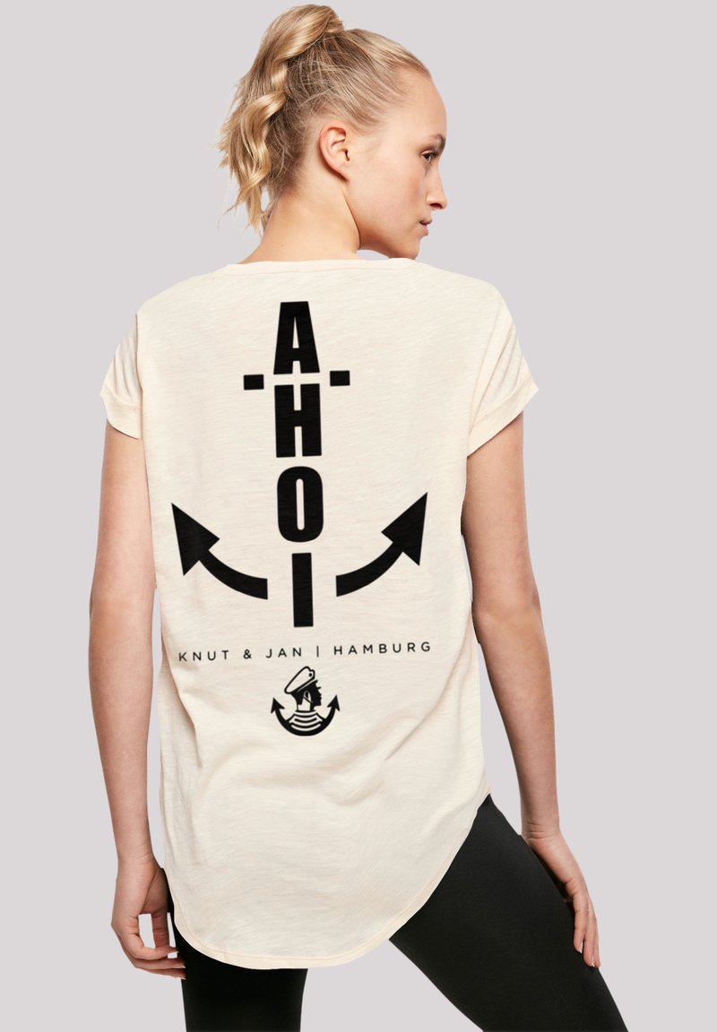 Cremefarbenes T-Shirt mit kurzen Ärmeln, das auf dem Rücken mit fettem schwarzen Text "AHOI" und Pfeilen sowie mit einer maritimen Grafik am unteren Ende versehen ist.