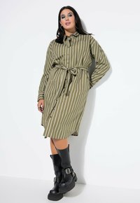 Olivgrünes gestreiftes Hemdkleid mit einer Knopfleiste und einem gebundenen Bund. Kombiniert mit hohen schwarzen Stiefeln mit Schnallenakzenten.