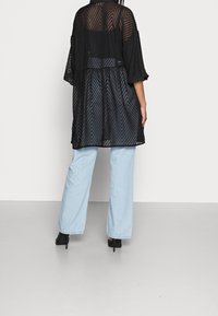 Sort gennemsigtig kimono med chevronmønster, løs pasform, vide ærmer og indsnævret talje. Båret sammen med lyseblå brede bukser og sorte hæle.