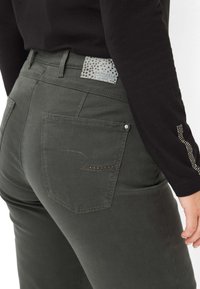 Pantaloni verdi scuri con una texture liscia, caratterizzati da un design con tasca posteriore, cuciture decorative e un'etichetta con motivi a stella.