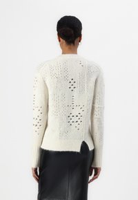 Krämfärgad stickad tröja med öppna mönster, ribbad mudd och sidoklipp. Mjuk textur, midjelängd och avslappnad passform.