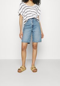 Mujer que lleva una blusa holgada a rayas en blanco y gris, pantalones cortos de mezclilla azul de tiro alto hasta la rodilla y sandalias beige tipo slip-on.