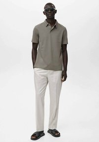 Cămașă polo gri cu guler, asociată cu pantaloni crem deschis. Modelul poartă ochelari de soare negri și sandale negre cu talpă joasă. Design simplu și modern.