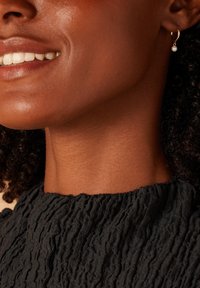 Boucle d'oreille en or avec une petite perle suspendue ; décolleté en tissu noir texturé ; teint de peau chaud ; gros plan sur la mâchoire et le sourire.