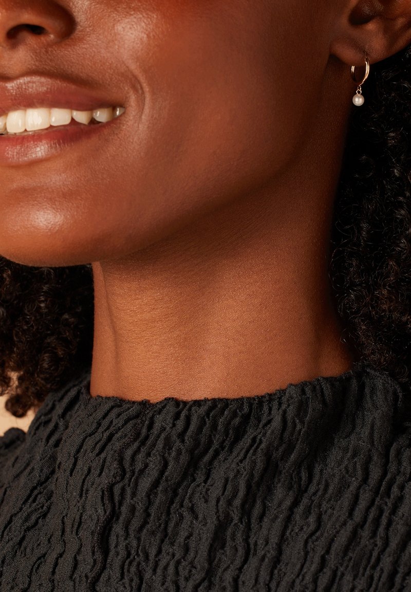 Boucle d'oreille en or avec une petite perle suspendue ; décolleté en tissu noir texturé ; teint de peau chaud ; gros plan sur la mâchoire et le sourire.