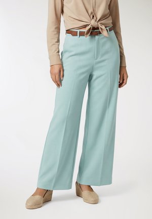 Donna che indossa una camicetta beige annodata, pantaloni larghi color acqua con cintura marrone e scarpe espadrillas beige con zeppa, in piedi davanti a uno sfondo semplice.