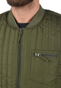 Bomber jakna u vojnicnozelenoj boji s kvadratnim uzorkom, koja ima prednji patentni zatvarač, rebrasti ovratnik i džep sa patentnim zatvaračem na lijevoj strani prsnog koša.