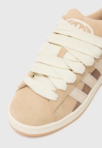 Sneaker with a beige suede upper, cream laces, and tan accents. Features a textured finish and three vertical stripes on the side.