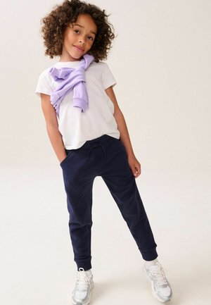 Kinderoutfit: weißes T-Shirt, marineblaue Jogginghose mit Taschen und leicht lila Pullover, der um die Schultern gebunden ist. Die Schuhe sind weiß mit silbernen Akzenten.