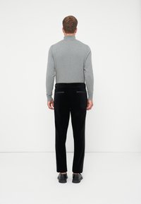 Muž stojí otočený chrbtom, oblečený v šedej košeli s dlhým rukávom, čiernych nohaviciach s zadnými vreckami a čiernych topánkach na pozadí jednoduchého bieleho pozadia.