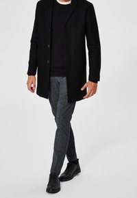 Manteau noir long sur un pull sombre, associé à un pantalon ajusté gris texturé et des chaussures noires à lacets. Design minimaliste avec des lignes épurées.