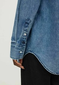 Šviesiai mėlynas džinsinis marškinėlių rankogalis su išlenktu apatine dalimi, dviem snapelių sagomis su baltomis detalėmis ir matoma siuvimo detalė.