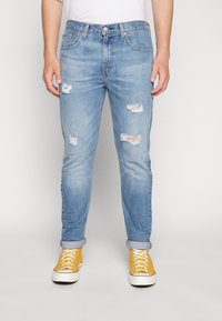 Hombre con jeans rotos de color azul claro y puños enrollados, usando zapatillas amarillas de caña alta, de pie contra un fondo blanco sencillo.