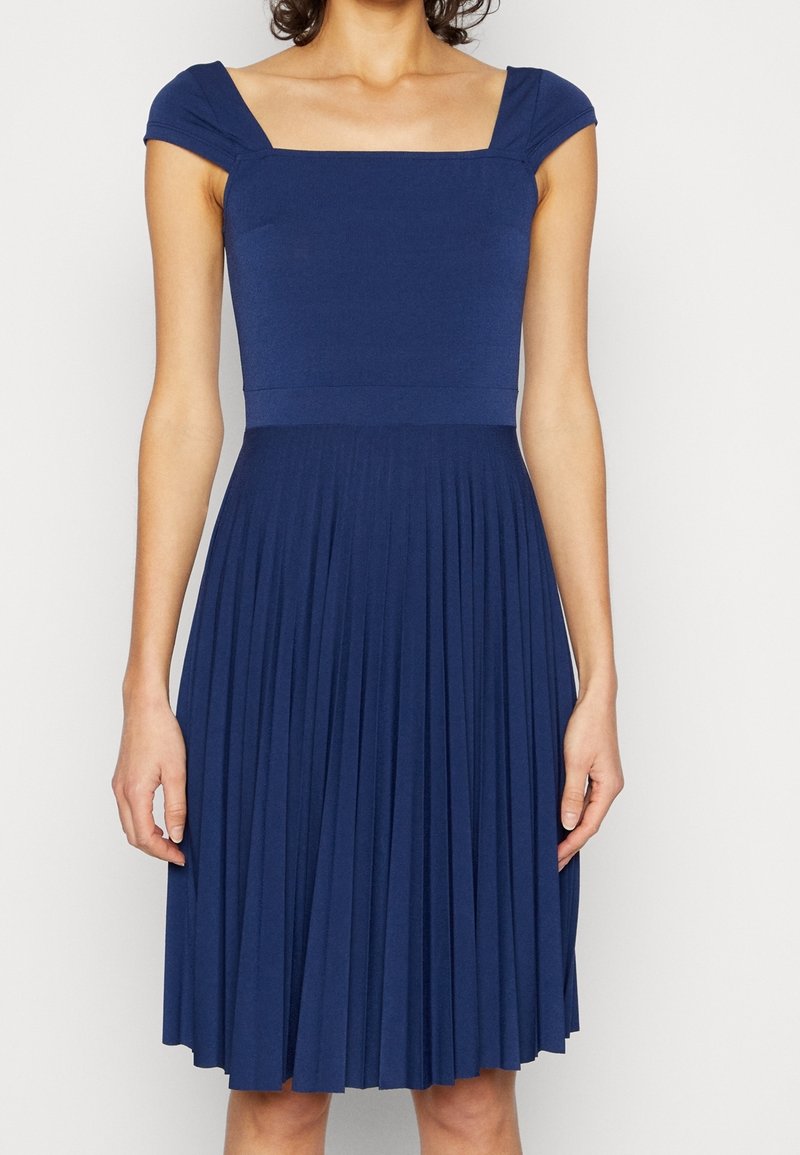 Femme portant une robe bleu marine sans manches avec un décolleté carré et une jupe plissée, debout devant un fond blanc.