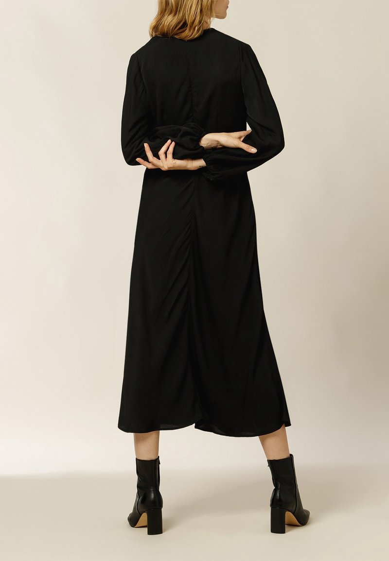 Femme aux cheveux blonds portant une robe midi noire à manches longues et des bottines noires à talons, debout, les mains croisées dans le dos.