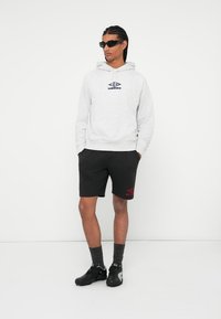 Uomo che indossa un hoodie grigio Umbro, pantaloni corti neri Umbro, scarpe sportive nere e occhiali da sole, in piedi contro uno sfondo bianco.