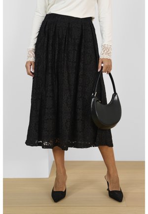 Kobieta w czarnej koronkowej spódnicy midi, białej bluzce z długim rękawem i koronkowymi mankietami, czarnych spiczastych szpilkach, trzymająca czarną skórzaną torebkę.