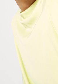 Top amarillo claro con escote drapeado, que presenta una tela suave y un ajuste relajado. El diseño enfatiza la simplicidad y la suavidad.