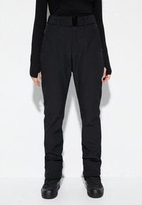 Persoon in zwarte high-waist broek met riem, langwerpig zwart shirt met duimgaten, en zwarte veterschoenen die tegen een witte muur staat.