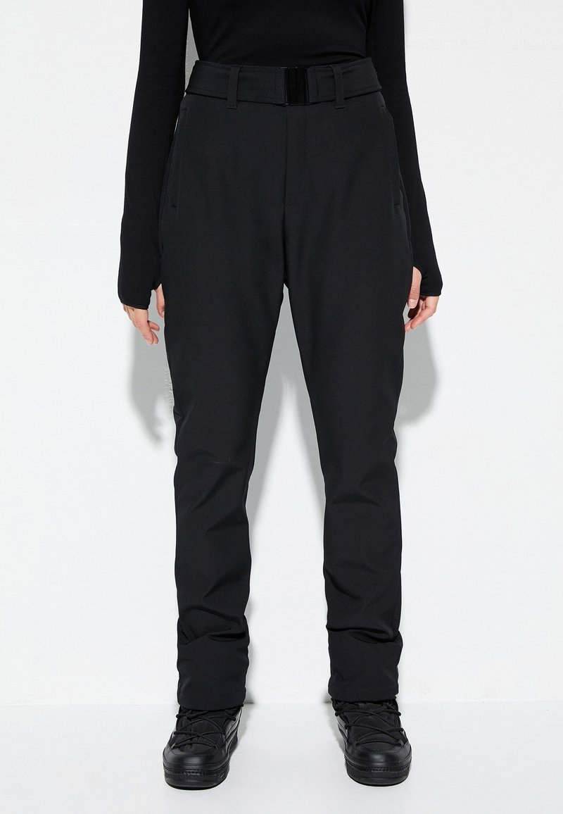 Persoon in zwarte high-waist broek met riem, langwerpig zwart shirt met duimgaten, en zwarte veterschoenen die tegen een witte muur staat.