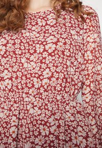 Abito rosso con motivo floreale bianco e arancione, vita arricciata e maniche lunghe leggermente voluminosi, indossato da una persona con capelli ricci.