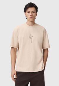 Camiseta de algodón de gran tamaño en color beige, con mangas cortas y cuello redondo, que presenta un gráfico de logo negro en el centro.