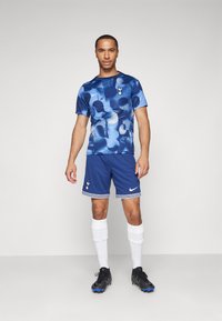 Blauw sportshirt met abstracte patronen, gecombineerd met donkerblauwe short. Witte logo-accenten, zwarte voetbalschoenen en witte sokken. Gladde textuur.