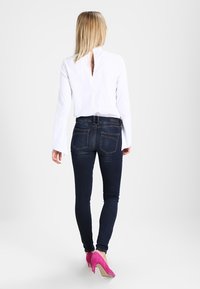 Blusa blanca de manga larga con diseño de espalda en V, combinada con jeans ajustados azul oscuro y zapatos de tacón rosa, vista desde atrás.