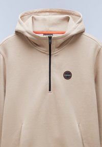 Sudadera beige con media cremallera, cremallera negra y textura sutil. Presenta un logo redondo en negro y naranja en el pecho.