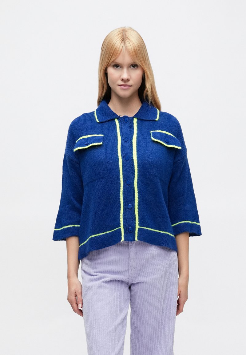 Niebieski dziany sweter z kołnierzem, krótkimi rękawami i wyraźnym neonowozielonym obszyciem. Posiada dwie przednie kieszenie i zapięcie na guziki.