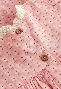 Robe en coton rose avec motif floral blanc, bordure en dentelle au niveau du col et boutons en bois. Présente des plis pour ajouter de la texture.